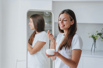 Satisfied caucasian young adult woman in white t-shirt stands at home holds can with creme trying new cosmetic product looks at camera smiles. Pretty Italian female at mirror uses skin care lotion.