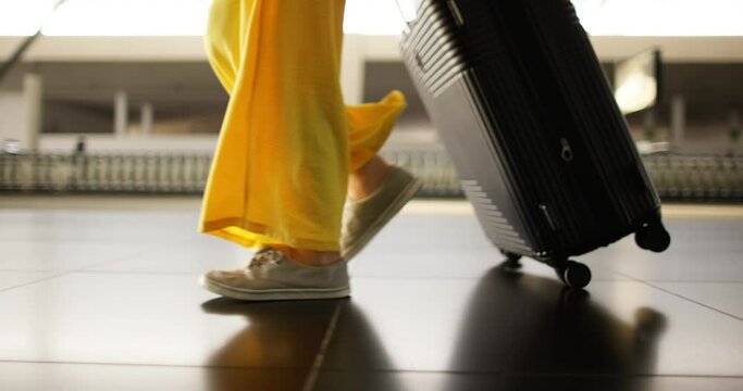Close up view of woman's legs in yellow pants and white sneakers. A woman slowly moves around the airport, waiting to board flight. A woman in departure area of airport walks, waiting to board plane.