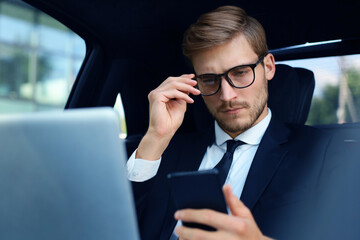 Handsome confident man in full suit looking at his smart phone while sitting in the car and using laptop.