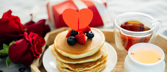 Pancakes with berries, roses flowers, cup of tea and candle in candlestick. Valentine's day breakfast concept. Banner for design, web site. Close-up - 561488896