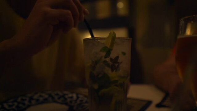 Woman hand mixing and drinking cocktail in glass with straw while sitting at table of cafe and talking with friend. Dating and meeting concept.