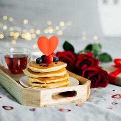 Valentine's day composition with breakfast, gift box and roses. Stack of delicious pancakes with berries and cup of hot moning tea on wooden tray