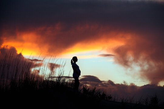 A pregnant woman is silhouetted against a colorful sunset while standing on a grassy hill.