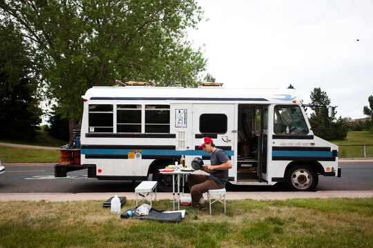A young man cooks dinner outside his short school bus which is converted to an RV in Denver, CO.