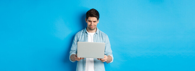 Disappointed handsome man looking at laptop screen and grimacing, judging something bad in...