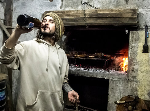 A Man Drinking A Beer While Cooking An Asado In Bariloche, Argentina