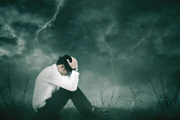 Stressed businessman sitting on the park with dried trees during thunder storm