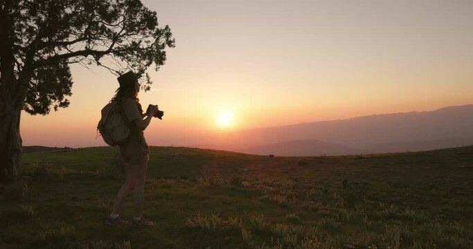 Silhouette of female journalist at summer sunset in mountains. Woman journalist takes beautiful photos of red sunset for her travel, adventure article. Hiking in mountains, professional photographer