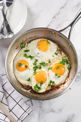 Four fried eggs with unbroken egg yolks, fresh green herbs on a metal dirty pan on a marble background, top view