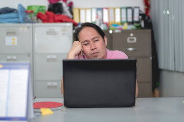 A bored asian man in his 40s watching a video on his laptop while sitting at his office desk.