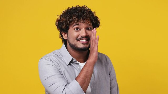 Mysterious fun happy young Indian man 20s wear casual grey shirt whispering gossip and tells secret behind HER hand sharing news isolated on plain yellow background studio. People lifestyle concept