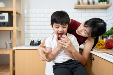 Asian little kid biting an apple with mother in the kitchen room. Attractive mom teach and support...