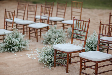 Fototapeta na wymiar Wedding wooden chairs decorated with flowers. Rustic aisle chairs standing on sand for ceremony on the beach. Natural, shabby, boho wedding decor