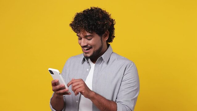 Young Indian man wear casual grey shirt hold use mobile cell phone typing browsing swipe chatting send sms doing online shopping order delivery to home booking tour isolated on plain yellow background
