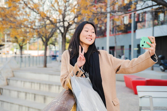 Cheerful young chinese or asian woman smiling taking a selfie portrait doing the peace sign to the cell phone after shopping and sharing in the social media with a smartphone app. High quality photo