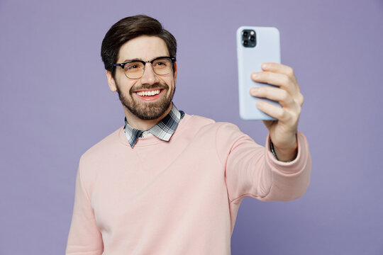 Young smiling IT man he wear casual clothes pink sweater glasses doing selfie shot on mobile cell phone post photo on social network isolated on plain pastel light purple background studio portrait.