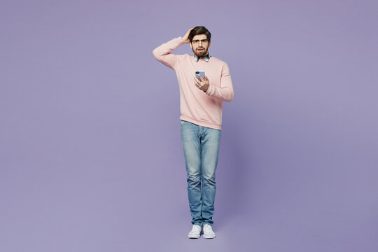 Full Body Scared Upset Young Caucasian IT Man He Wear Casual Clothes Pink Sweater Glasses Hold Head Use Mobile Cell Phone Look Camera Isolated On Plain Pastel Light Purple Background Studio Portrait.