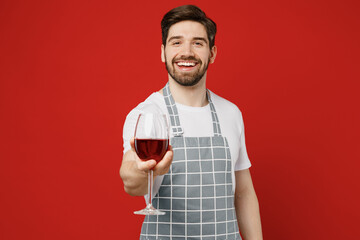 Young smiling sommelier happy male housewife housekeeper chef cook baker man wear grey apron hold in hand give glass of wine look camera isolated on plain red background studio. Cooking food concept.
