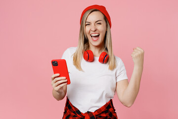 Young overjoyed excited happy satisfied positive woman wear white t-shirt red hat hold in hand use mobile cell phone do winner gesture isolated on plain pastel light pink background studio portrait.