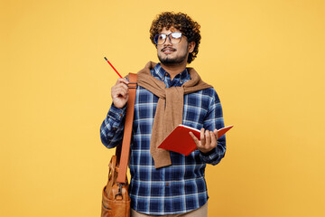 Young teen Indian boy IT student wear casual clothes glasses bag write in notebook point finger pencil aside on area isolated on plain yellow color background High school university college concept.