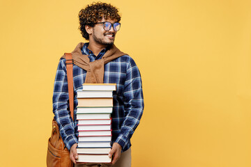 Fototapeta Young teen Indian boy IT student wear casual clothes shirt glasses bag hold in hands pile of books look aside on area isolated on plain yellow color background High school university college concept. obraz