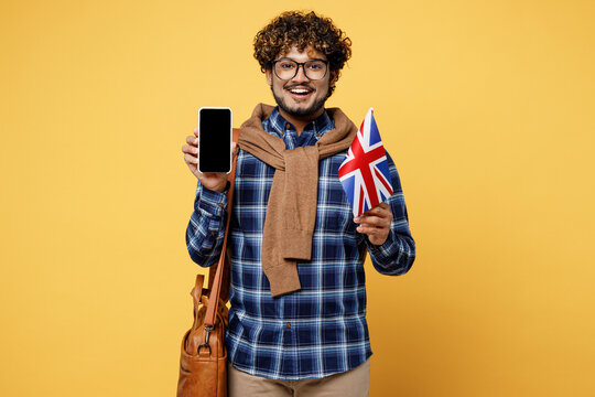 Young teen Indian boy IT student wear casual clothes glasses bag hold mobile cell phone blank screen area British flag isolated on plain yellow color background High school university college concept