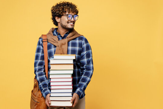 Young teen Indian boy IT student wear casual clothes shirt glasses bag hold in hands pile of books look aside on area isolated on plain yellow color background High school university college concept.