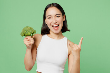 Young woman wears white clothes hold in hand broccoli vegetable showing horns up gesture isolated on plain pastel light green background. Proper nutrition healthy fast food unhealthy choice concept.