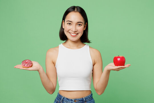 Young smiling fun woman wear white clothes hold in hand red apple pink donut look camera isolated on plain pastel light green background. Proper nutrition healthy fast food unhealthy choice concept.