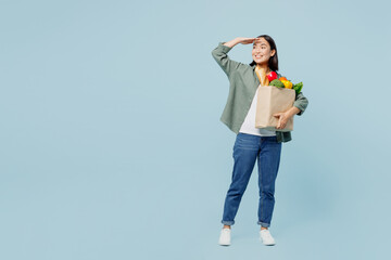Full body young woman wear casual clothes hold brown paper bag with food products look far away for taxi isolated on plain blue background studio portrait. Delivery service from shop or restaurant.