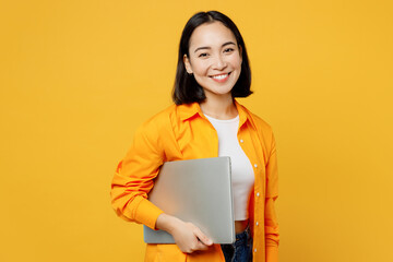 Young IT woman wears summer casual clothes hold closed laptop pc computer isolated on plain yellow...