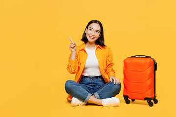 Young woman in summer casual clothes sit near suitcase point aside on area isolated on plain yellow...