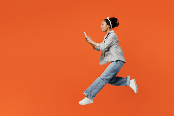 Fototapeta na wymiar Full body side profile view excited young woman of African American ethnicity she wears grey shirt headband jump high hold in hand use mobile cell phone run fast isolated on plain orange background.