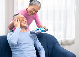 Senior woman surprises her husband with a gift box. Happy old Asian couple enjoy celebration with valentine present. Romance elderly husband and wife giving gift in anniversary marriage celebration.