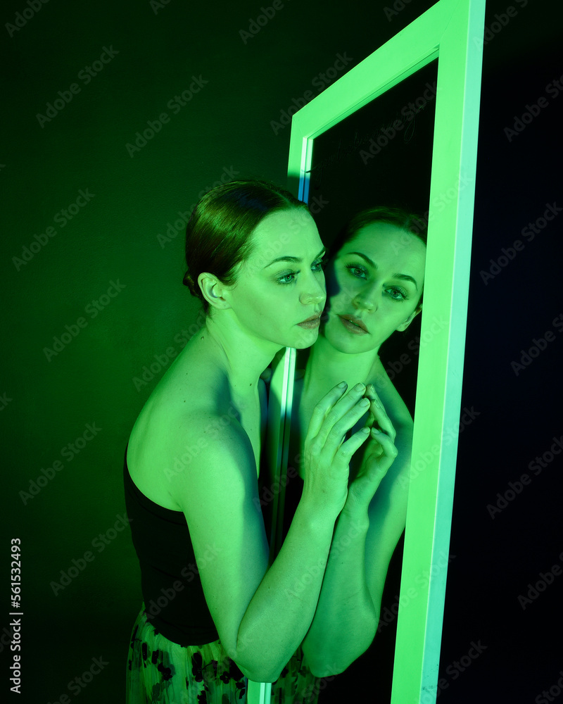 Poster Close up portrait of beautiful girl looking into mirror, self reflection with colourful studio lighting.