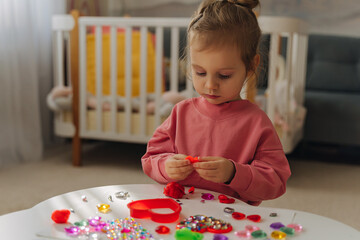 A little girl playing with red heart from play dough for modeling and  decorations from crystal rhinestones and shiny stones. Toddlers crafts for Valentine's Day. Holiday Art Activity for Kids.