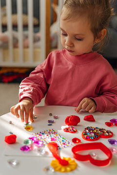 A little girl playing with red heart from play dough for modeling and  decorations from crystal rhinestones and shiny stones. Toddlers crafts for Valentine's Day. Holiday Art Activity for Kids.