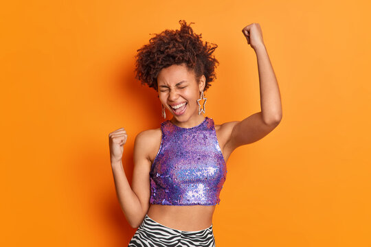 Studio shot of optimistic lucky woman clenches fists raises arms up celebrates success exclaims loudly feels like winner has slim figure dressed in cropped purple top isolated over orange background