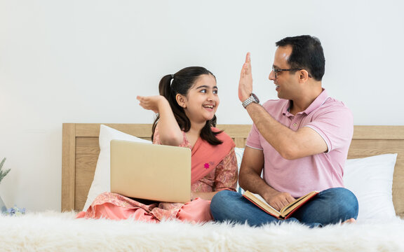 Indian happy father and daughter sitting on bed at cozy home, smiling with success, happiness, teaching, doing homework, playing, using laptop, high five. Education, Technology, Activity Concept.