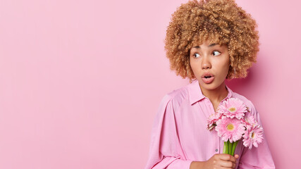 Indoor shot of good looking woman with curly hair holds nice bouquet receieves flowers on special occasion dressed in fashionable shirt isolated over pink background copy space for your text