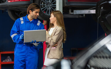 Handsome male mechanic wearing uniform, using laptop, checking or inspecting for fix, repair car or automobile components, talking with customer, working in car maintenance service center or shop.