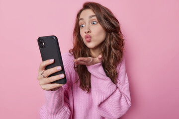 Fototapeta premium Romantic beautiful dark haired young woman blows air kiss at camera has video chat with boyfriend expresses love keeps lips rounded dressed in loose knitted jumper isolated over pink background