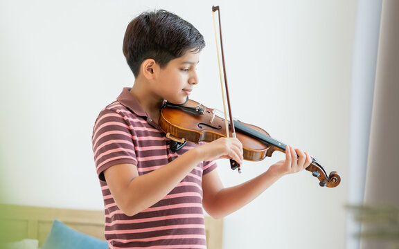 Indian handsome teenage boy, smiling, playing, practicing violin musical instrument with happiness in bedroom at cozy home in leisure time, smiling, having copy space. Education, Lifestyle, Concept.
