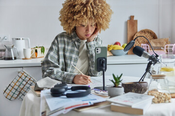 Fototapeta na wymiar Focused curly haired female student has online courses learns foreign languages writes down important information in notebook poses against home interior. Remote studying and education concept
