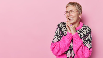 Horizontal shot of happy sincere short haired woman keeps hands together near face smiles toothily wears big optical spectacles and loose warm jumper isolated over pink background copy space for promo © WHstudio Leushin N