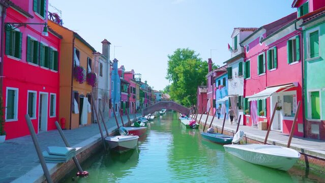 Bright multi-colored semi-detached houses stand on water channel banks with moored boats under bright sun rays and clear blue sky Burano
