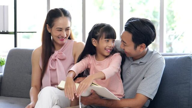 4k Video of father, mother and daughter sitting on sofa reading a book and playing educational games in living room, they smiling and laughing together happily on vacation. family life is happiness.