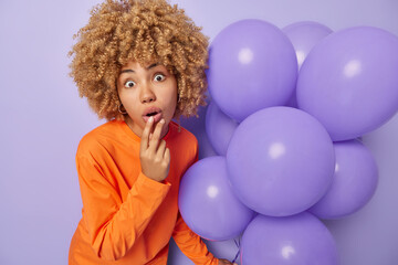 Fototapeta na wymiar Surprised woman keeps mouth opened jaw dropped reacts to shocking news wears orange long sleeved jumper holds bunch of inflated balloons celebrates special occasion holds bunch of inflated balloons