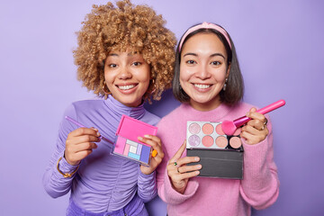 Indoor shot of cheerful biracial women hold palettes of colorful eyeshadow and cosmetic brushes apply makeup smile toothily prepare for party undergo beauty procedures isolated over purple background