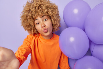 Fototapeta na wymiar Romantic woman makes selfie poses near inflated balloons keeps lips folded sends air kiss at camera dressed in orange sweater celebrates anniversary isolated on purple background. People and holidays